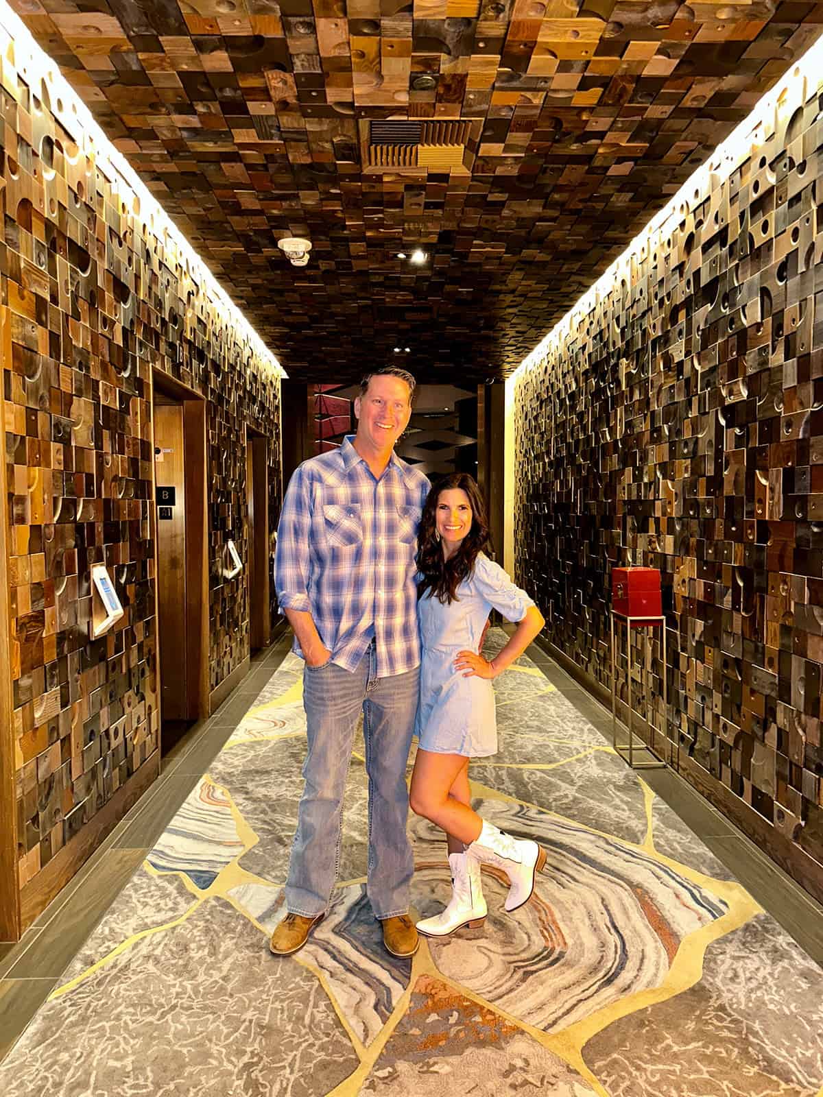 A man in plaid and a woman in a blue romper with white boots on standing in a hallway of bronze tiles.